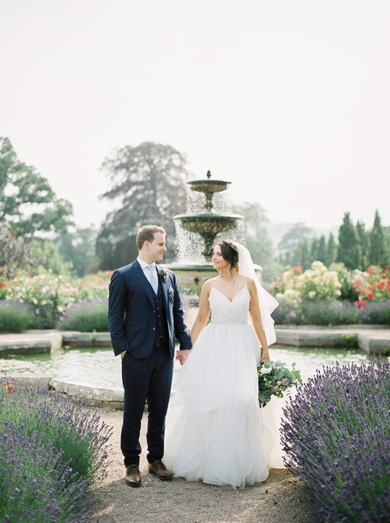 Bride and groom standing by fountain in rose gardens at country house in Devon