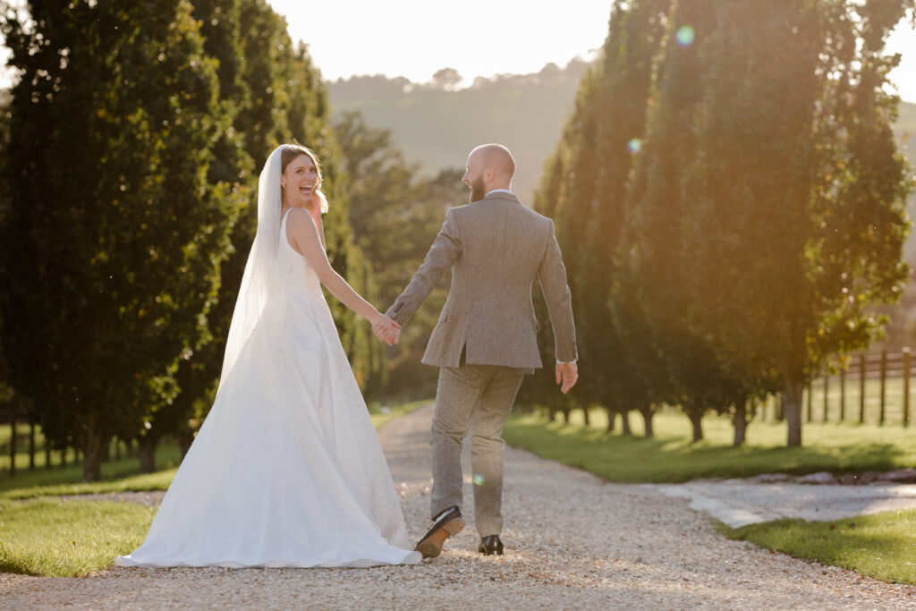 Bride and groom walking down tree lined drive.