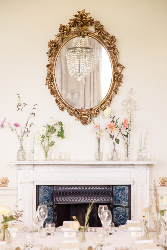 Gold mirror with fireplace underneath. Fireplace has vases of pink and white flowers on the mantlepiece above it.