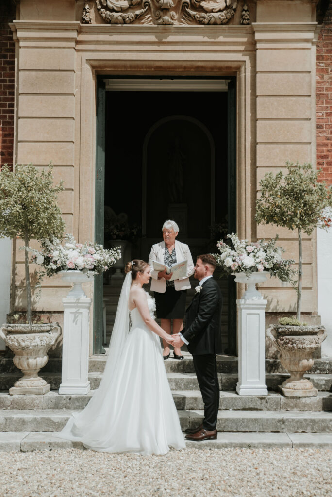 Bride and groom holding hands at the altar outside brick English country house.