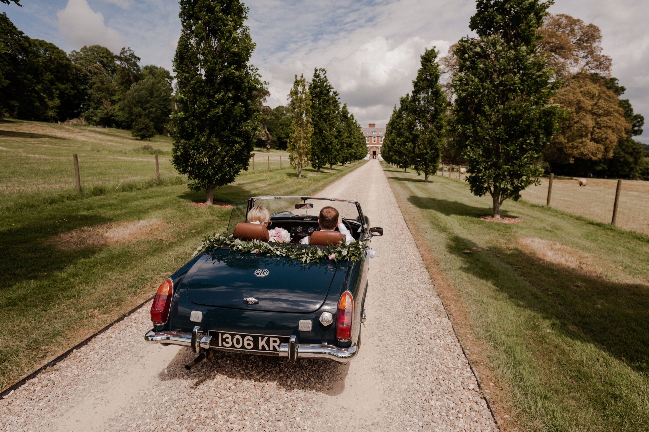 Bride and groom driving an open top car driving down long driveway.