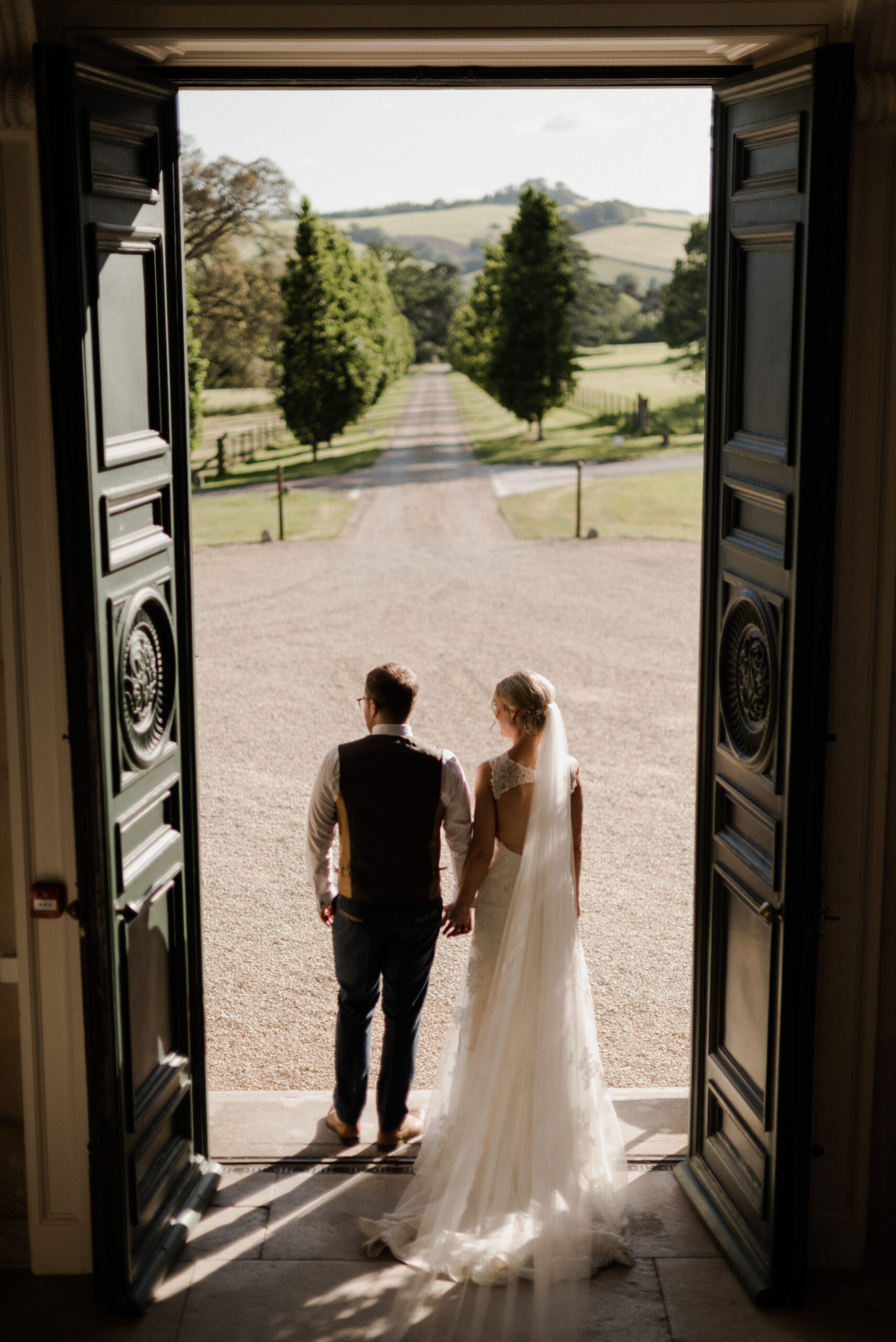 Bride and groom standing by front doors looking down driveway.