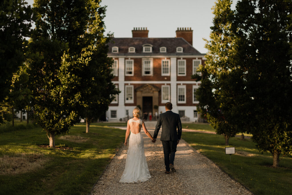 Bride and groom holding hands, walking towards English country house.
