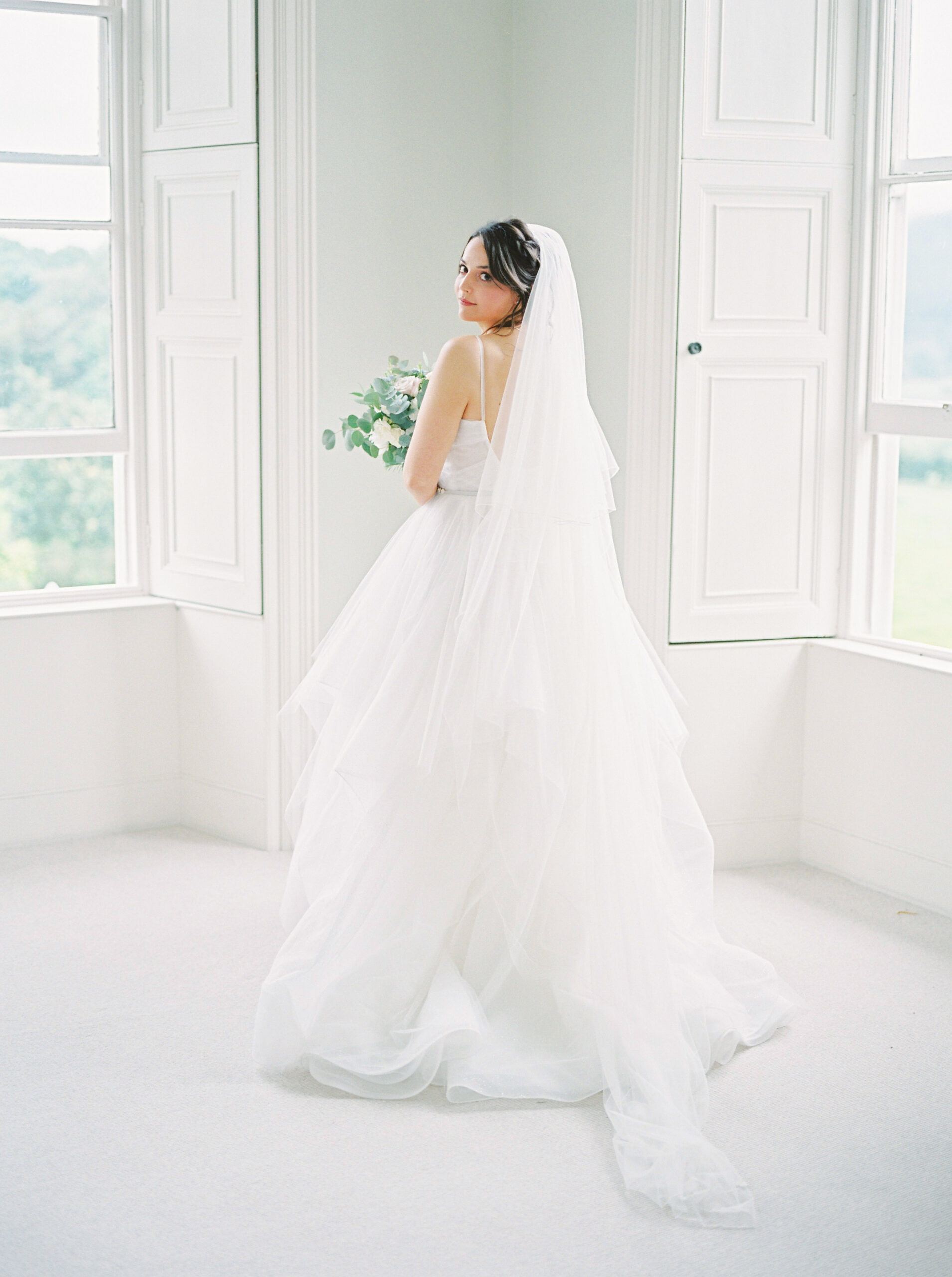 Bride standing in bedroom in wedding dress, holding flowers.