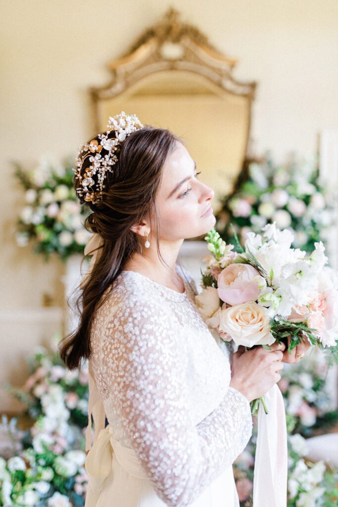 Bride holding bouquet, with large mirror and flowers behind her.