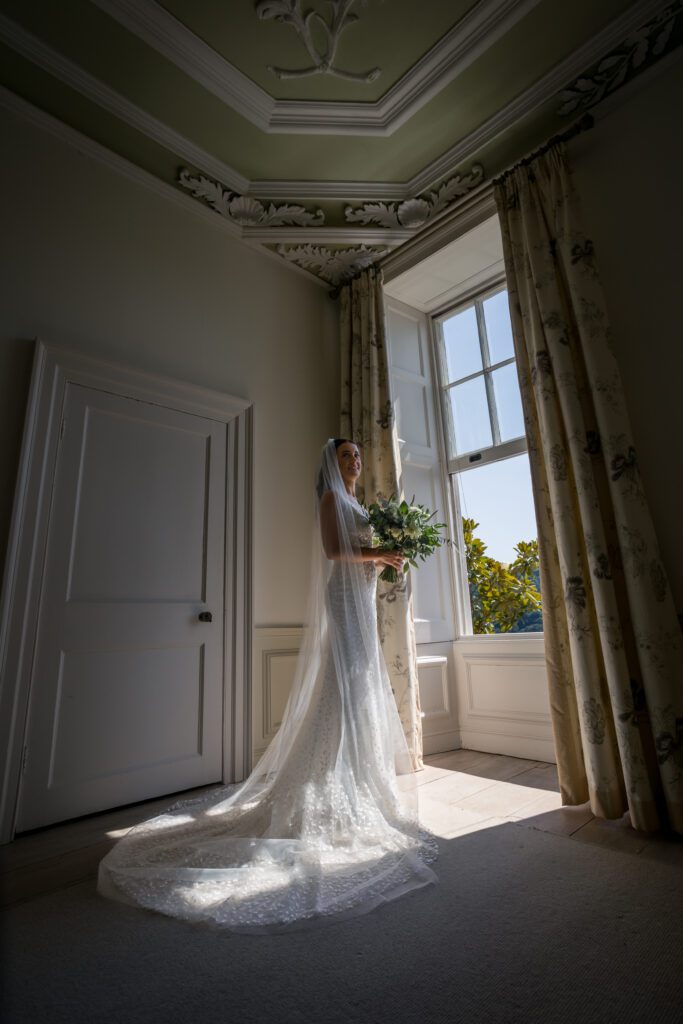 A bride stands elegantly by a window in her wedding dress during bridal prep, on the morning of her country house wedding.