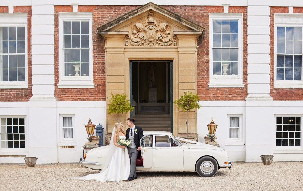 Bride and groom standing in front of their dream wedding venue with a classic white bridal car.